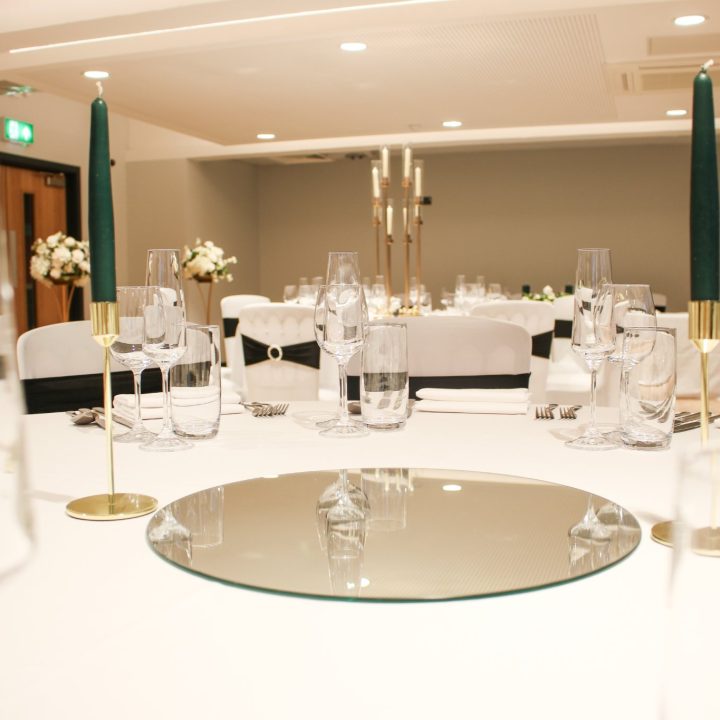 A round table set for an event, with a white tablecloth, elegant glassware, a mirror centrepiece.