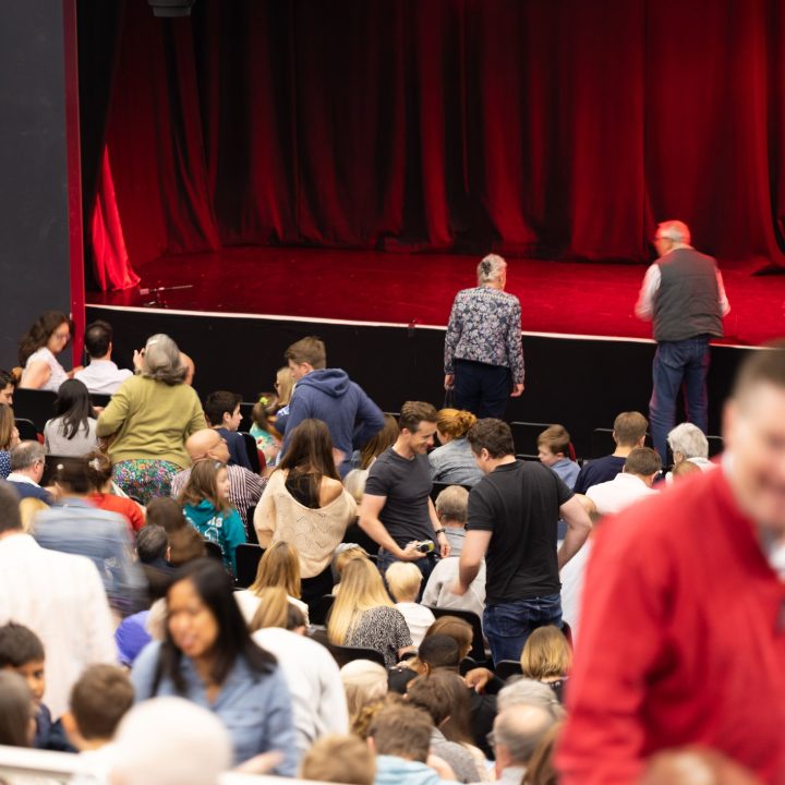 A crowded theater audience is seated in anticipation. Two people are seen walking towards the stage.