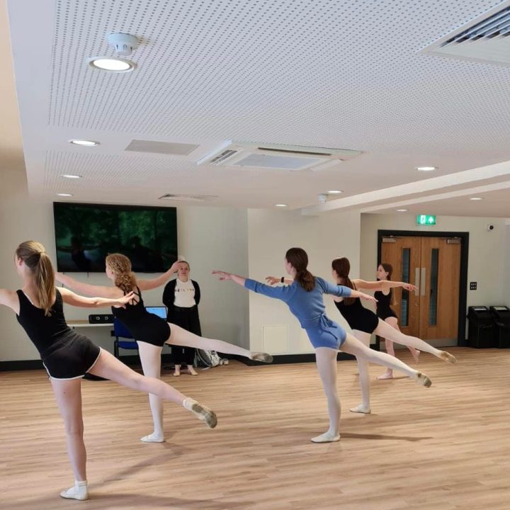 A group of ballet dancers in a studio practice an arabesque pose, with one leg extended behind them.