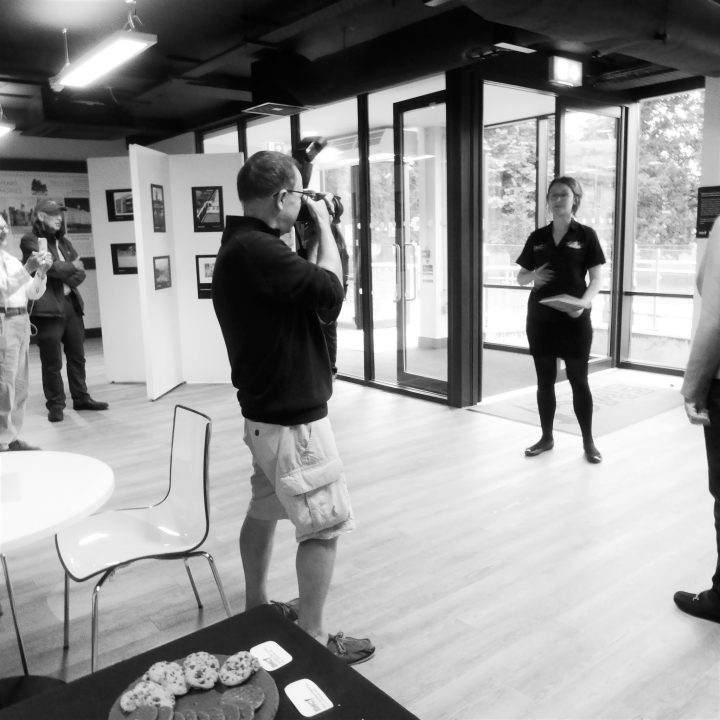 A person is taking a photo of another individual speaking to a small group in a gallery setting.