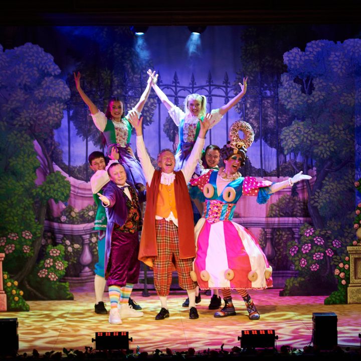 A lively stage production with vibrant costumes features six performers standing.