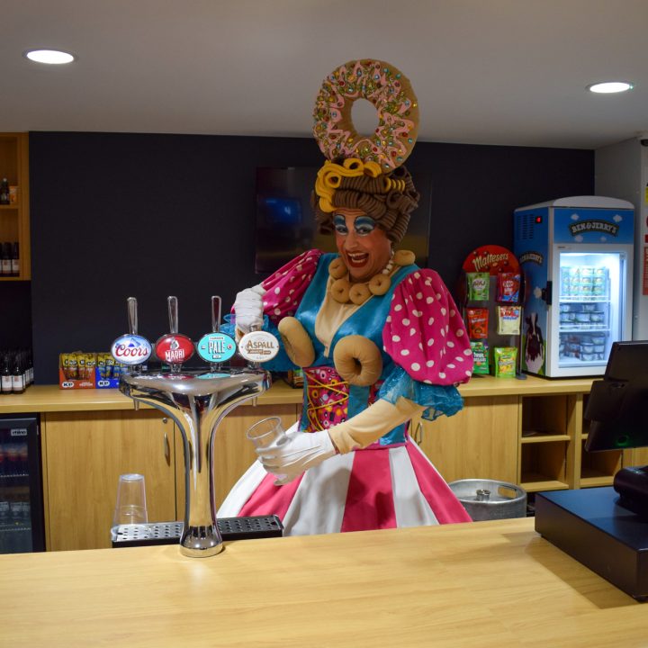 A person dressed in a colourful, whimsical costume with large pastries on their head.