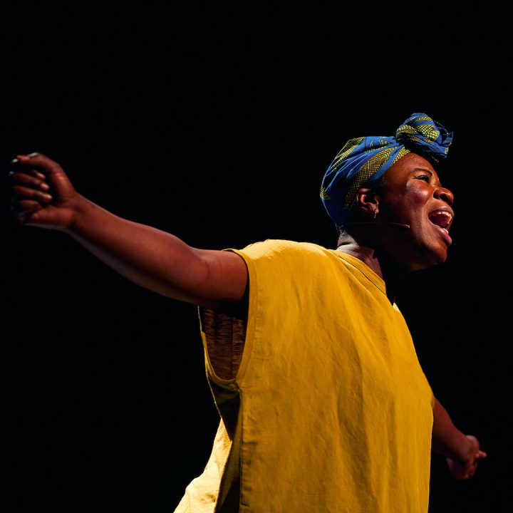 A person joyfully singing with outstretched arms, wearing a yellow garment.