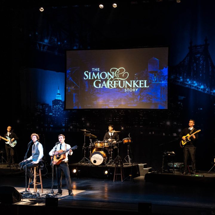 A band performs on stage with a backdrop reading "The Simon & Garfunkel Story."