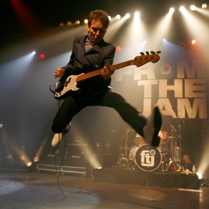 A musician dressed in a dark suit jumps in mid-air while playing a guitar.
