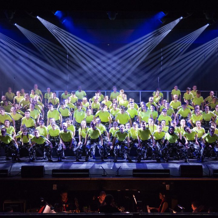 A large choir wearing matching bright green shirts performs on stage under a series of spotlights.