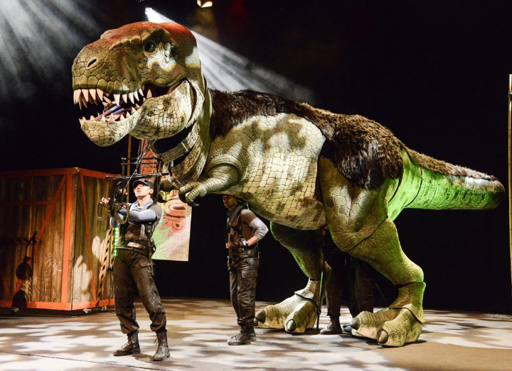 A large, realistic T-rex puppet is featured on Alban Arena stage.