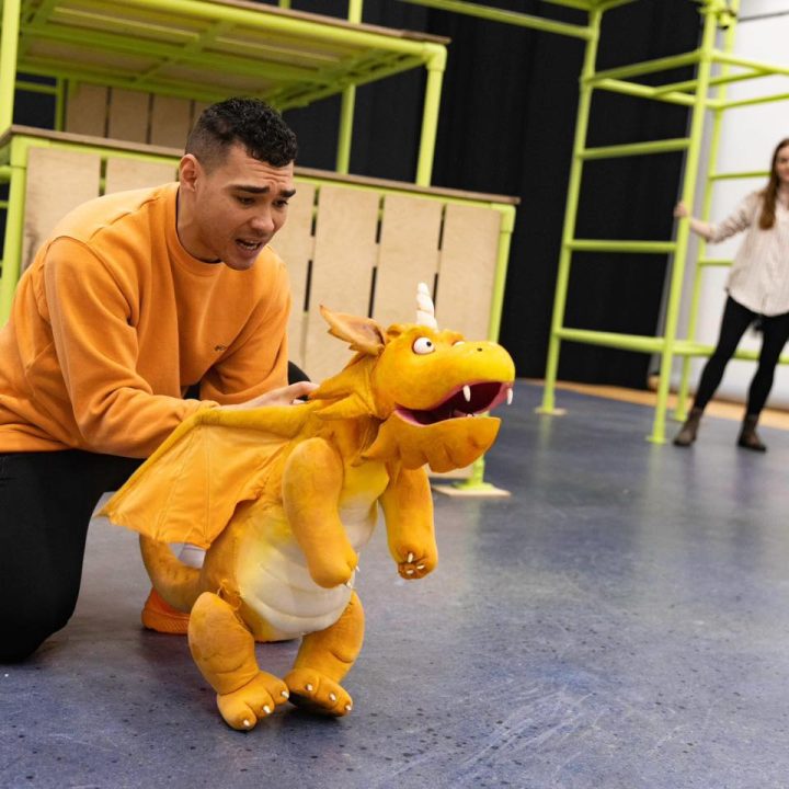 A person kneels on the floor while holding a small, orange dragon puppet.