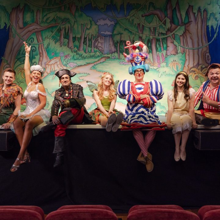 A group of people in colourful, whimsical costumes sit and stand on the edge of a stage.
