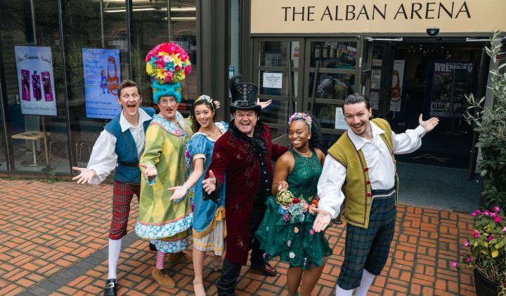 A group of six people in colourful, theatrical costumes stand happily with arms open.