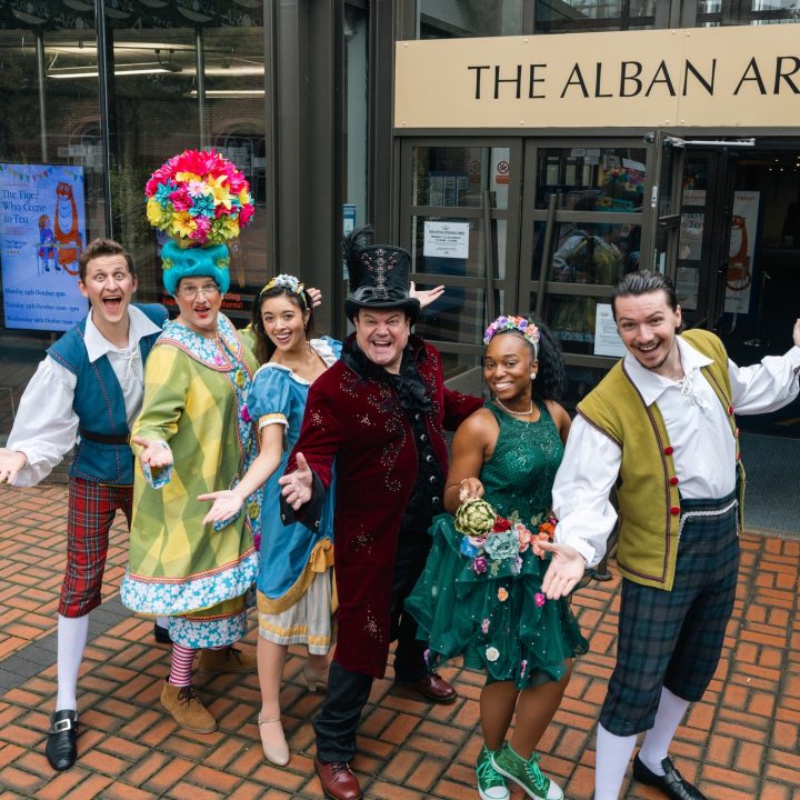 A group of six people in colourful, theatrical costumes stand happily with arms open.