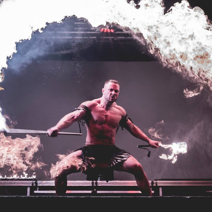 A muscular man performing a fire dance stands in a low stance holding a flaming staff.
