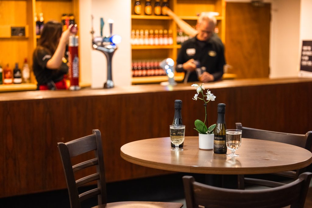 Table with wine bottles and glasses in a bar, as two people handle pre-ordering options nearby.