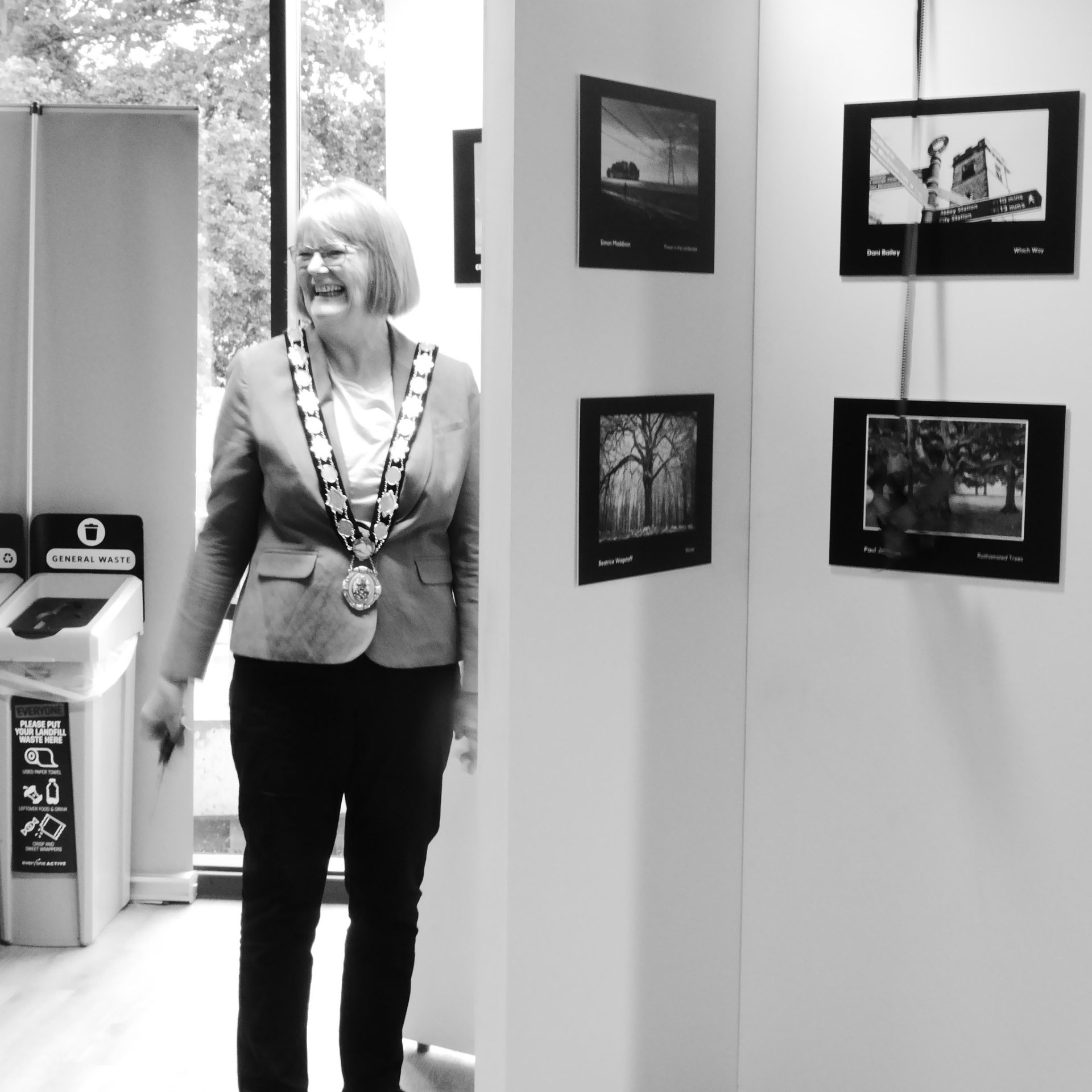 A smiling person with a chain stands near framed photos in Harpenden's history gallery.
