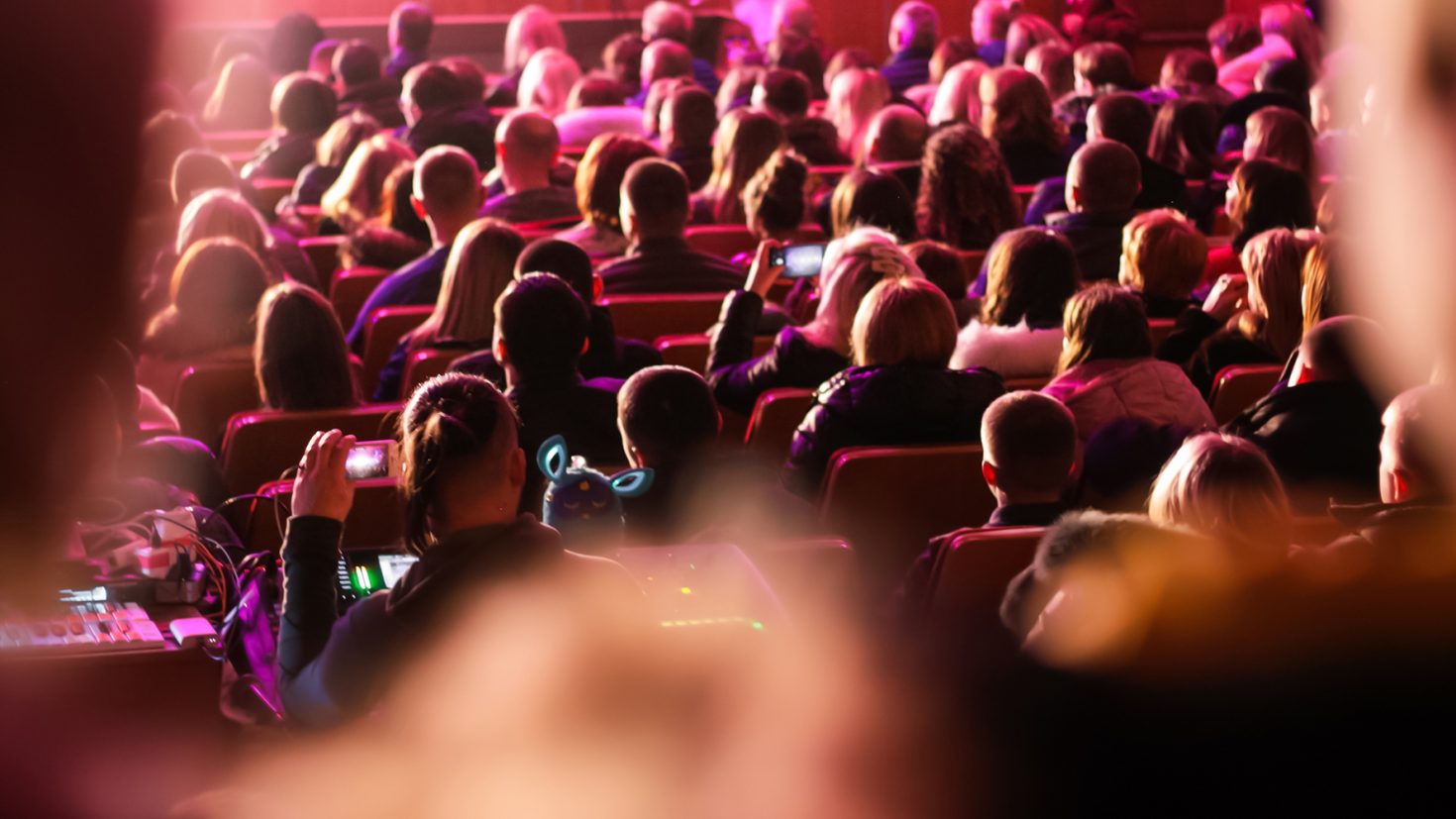 Audience seated in a theatre, facing the stage, under pink lighting.
