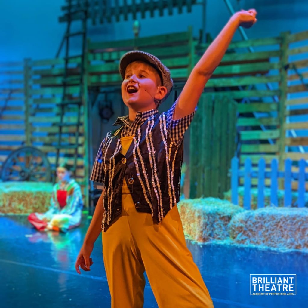 Child shines on stage in bright costume with farm backdrop; another sits.